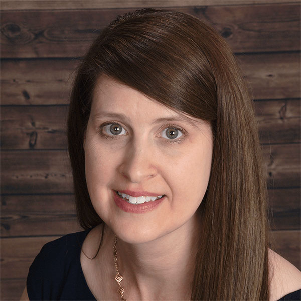The image shows a woman with long brown hair, wearing a dark top, smiling slightly and looking directly at the camera. She has a necklace and appears to be indoors with a wooden background.