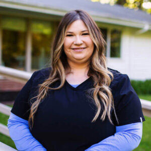 A woman in a black shirt and blue jeans stands confidently outdoors, smiling at the camera.