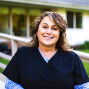 The image shows a woman wearing scrubs and standing outdoors, with a clear sky in the background.