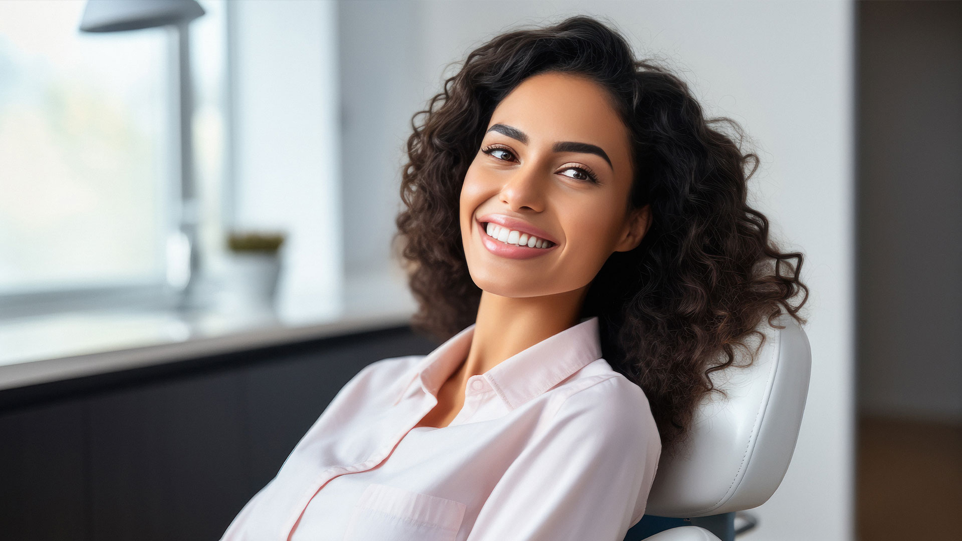 A woman with curly hair, smiling at the camera, seated in a modern office environment.