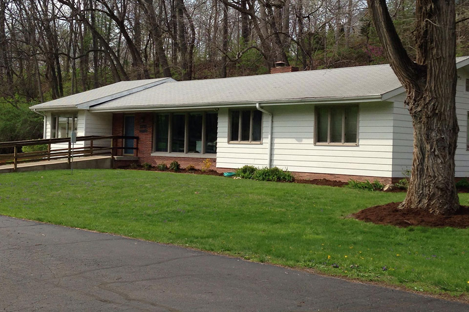 The image shows a single-story house with a white exterior, a gray roof, and a brick chimney. It has a covered porch with a railing and steps leading up to it. There is a driveway with a concrete surface that leads to the garage door on the left side of the house. The front yard is landscaped with grass and a few small trees or shrubs. The sky appears overcast, suggesting an overcast day.