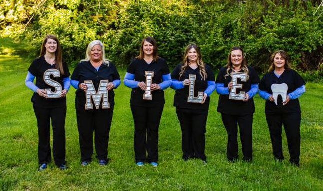 The image is a group photograph of nine individuals wearing blue scrubs, each holding a letter that spells out smile with a large white smiley face in the center. They are standing on grass with trees and foliage behind them, under a partly cloudy sky.
