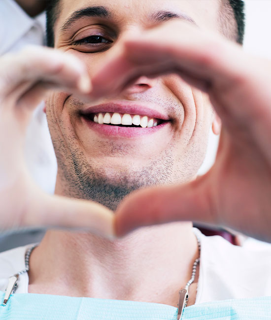 The image shows a smiling man in a dental chair, holding up two heart-shaped hands with his eyes closed. He appears to be in a dental office setting, wearing a medical mask and face shield, and there s visible dental equipment around him.