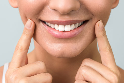 The image shows a close-up of a person s face, focusing on their smiling mouth and the application of what appears to be a skincare product, with the individual using both hands to spread it across their cheeks.