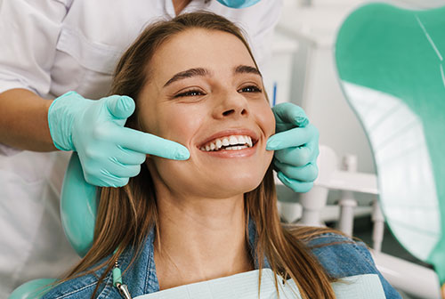 The image shows a woman sitting in a dental chair with her eyes closed, smiling broadly, while undergoing dental treatment a dentist is adjusting her teeth with a smile on their face.
