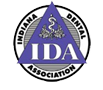 The image is a logo featuring the acronym IDA in large, bold letters, with the word Association directly below it. Above the acronym, there is an emblem that includes a stylized depiction of a dental implant and a circular design resembling a shield or badge. The logo has a central, rectangular shape with rounded corners, set against a background that transitions from white at the top to a darker shade towards the bottom.