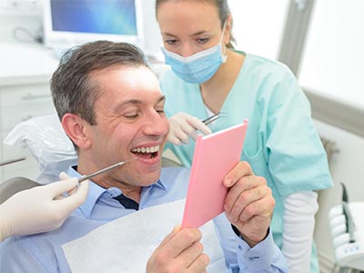 The image shows a man in a dental office, holding up a pink card or sign, with a smile on his face and surrounded by dental equipment. A woman wearing a surgical mask is standing behind him, possibly a dental professional.
