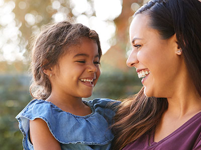 The image depicts a woman and a young child, both smiling, with the woman holding the child. They are outdoors during daylight, possibly in a park or similar setting.