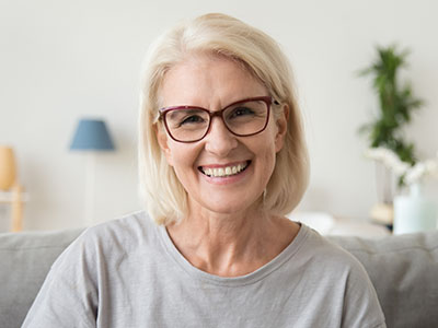 The image shows a woman with short blonde hair, wearing glasses and a light-colored top, seated indoors with a smile on her face.