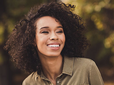 The image shows a person with curly hair smiling at the camera. They are wearing a dark top and have a relaxed posture.